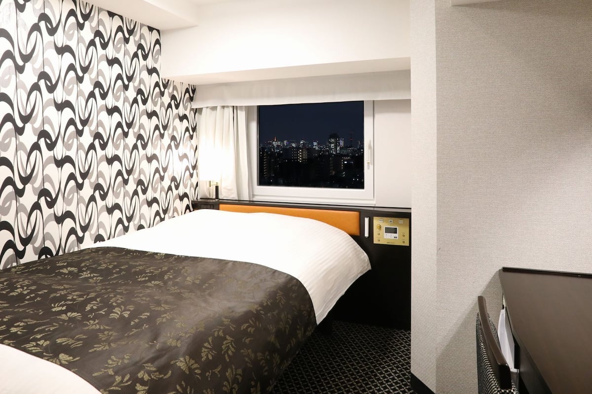 A hotel room with a bed, a window with a city view, a bedside table, and a mirror.