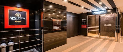 Entrada d'hotel amb una recepció, un senyal que indica l'escriptori d'informació i una porta amb la inscripció 'APA HOTEL'.