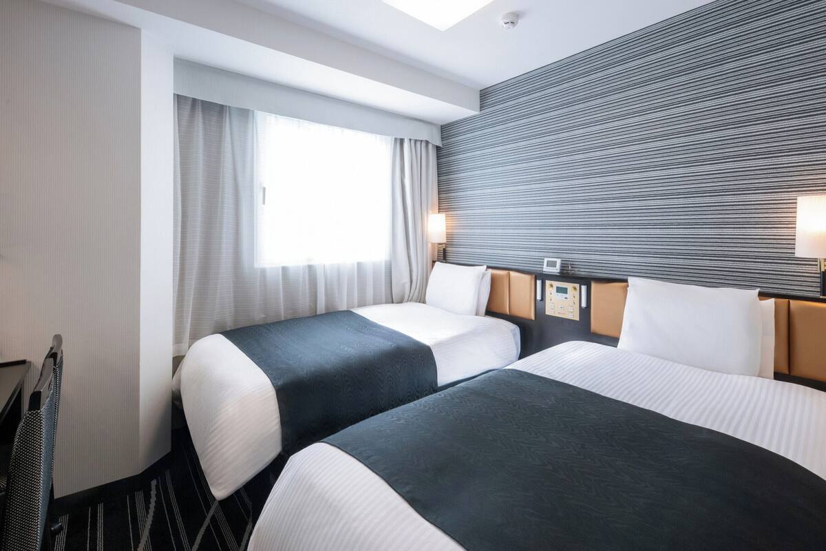 Ein Hotelzimmer mit zwei Betten, einem großen Fenster mit Vorhängen, einem Kopfteil mit Bedienfeld und einer Wand mit Strukturmuster.