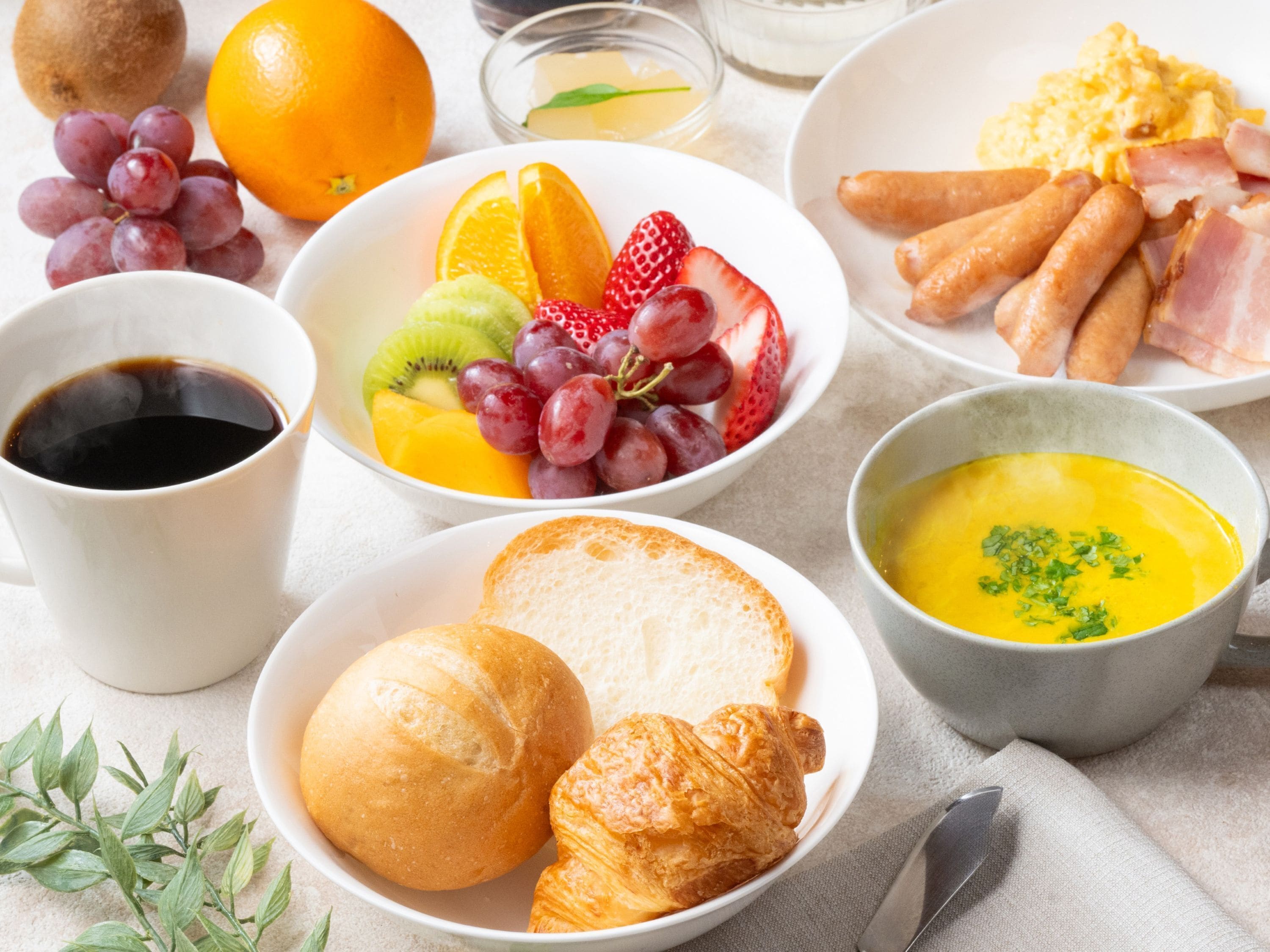 Ein Frühstückstisch mit Croissants, Brot, Obst, Rühreiern, Würstchen und einer Schüssel Suppe.