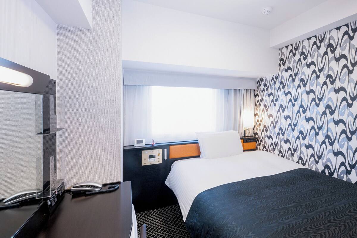 Ein Hotelzimmer mit einem Bett, einem Schreibtisch mit Computer, einem Telefon und einer Bedienkonsole.