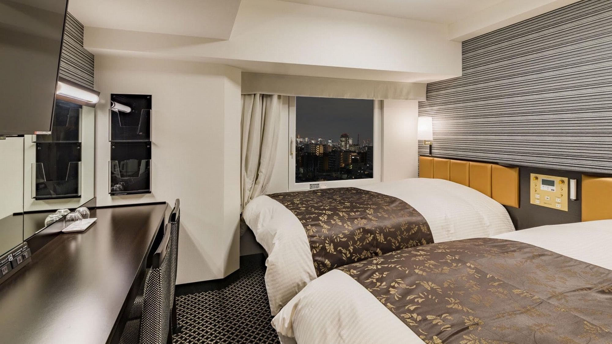 A hotel room with two beds, a desk, a window with a city view, and a control panel on the headboard.