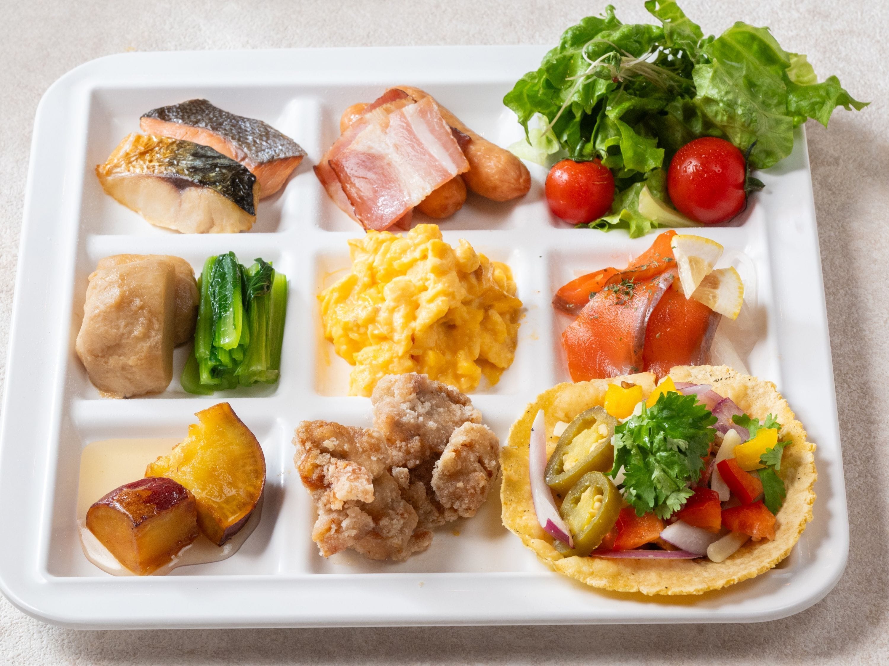 A white rectangular tray with various food items including fish, vegetables, scrambled eggs, and a taco.