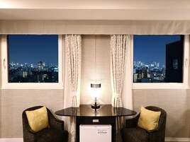 Habitación de hotel con vista a la ciudad, una mesa redonda de vidrio, dos sillones estampados y un refrigerador blanco pequeño.