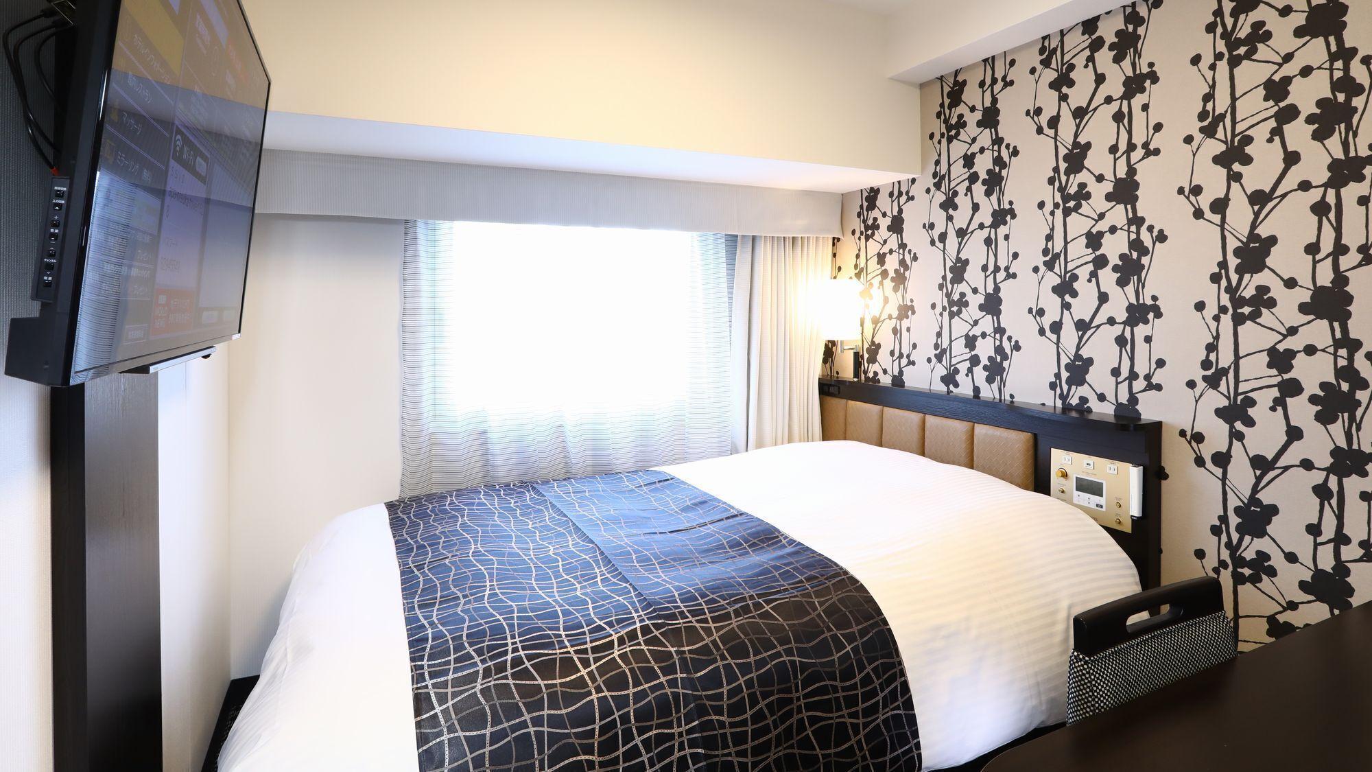 A hotel room with a bed, a television, a window with curtains, a wall with a floral pattern, and a bedside table.