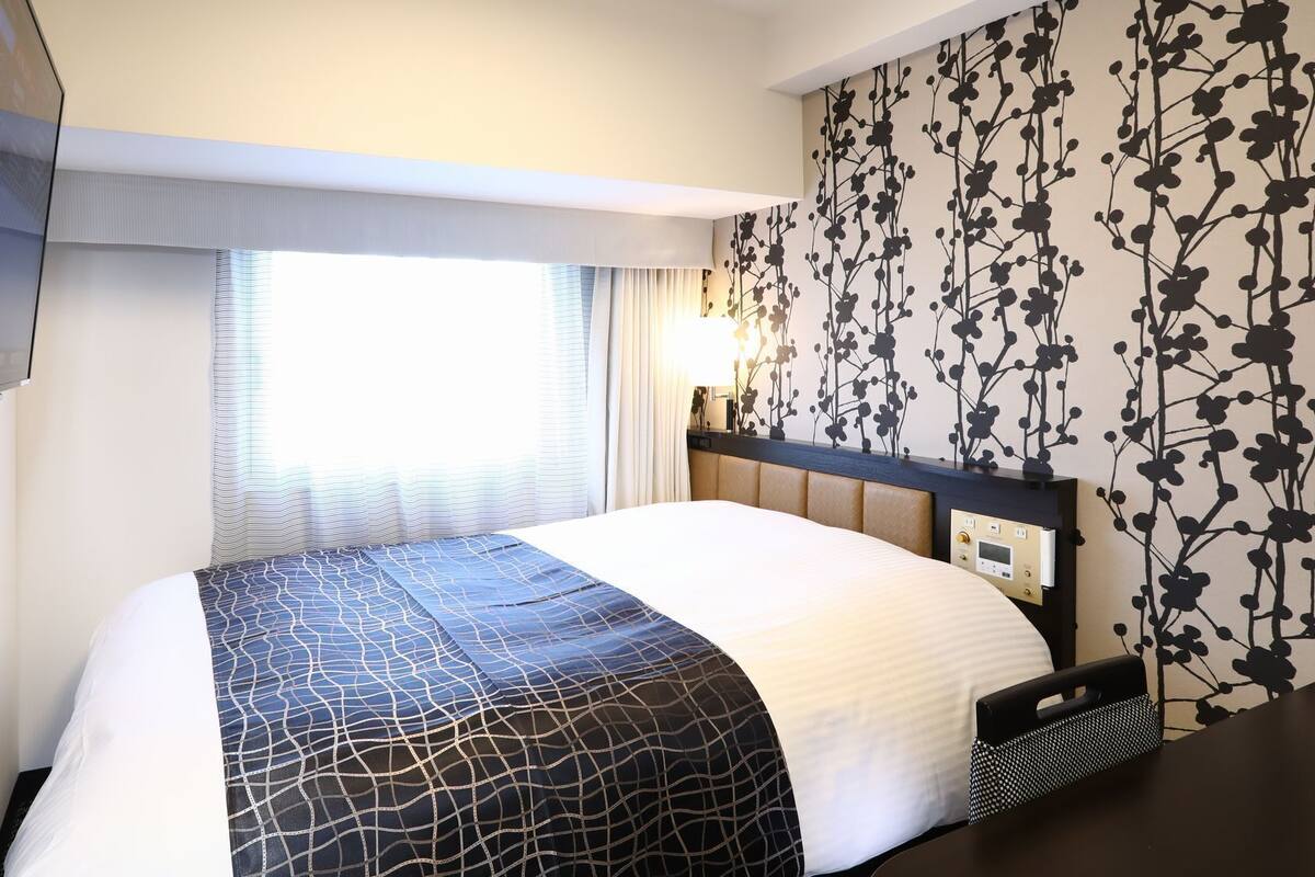 Une chambre d’hôtel comprenant un lit, une télévision, une fenêtre avec des rideaux, un mur orné d’un motif floral et une table de chevet.