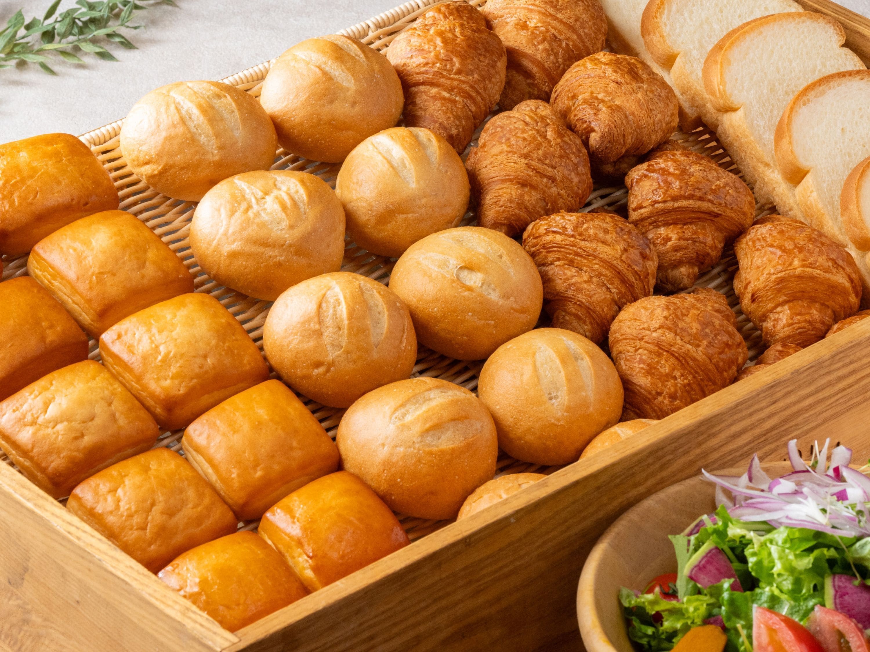 A wooden basket filled with various types of bread, including baguettes and croissants, alongside a bowl of salad.