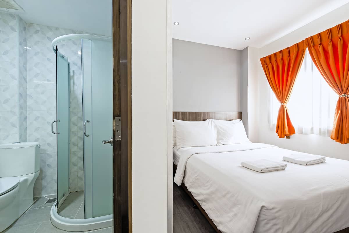 A hotel room with a glass-enclosed shower, a bed with white linens, and orange curtains.