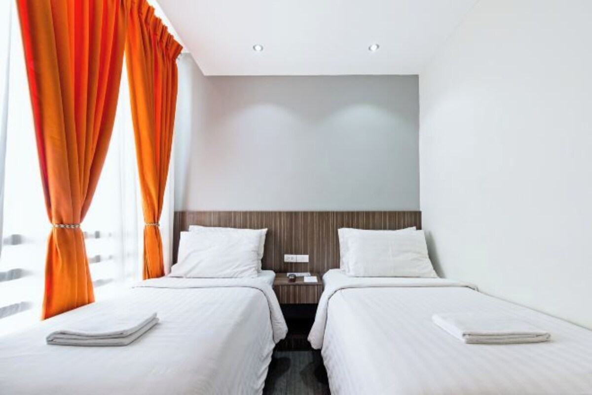 Habitación de hotel con dos camas, cabecero de madera, ropa de cama blanca y cortinas naranjas.