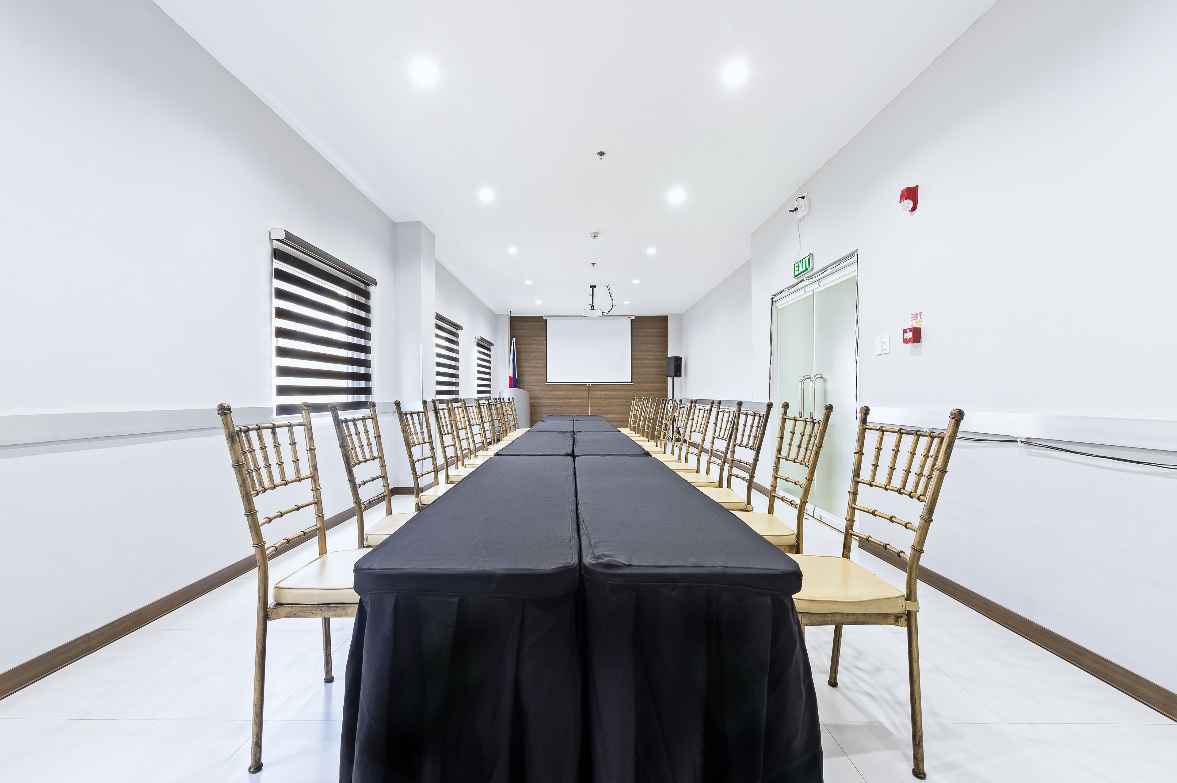 Un salón de conferencias con una mesa larga, sillas, una pantalla de proyector y una puerta con letrero de salida.