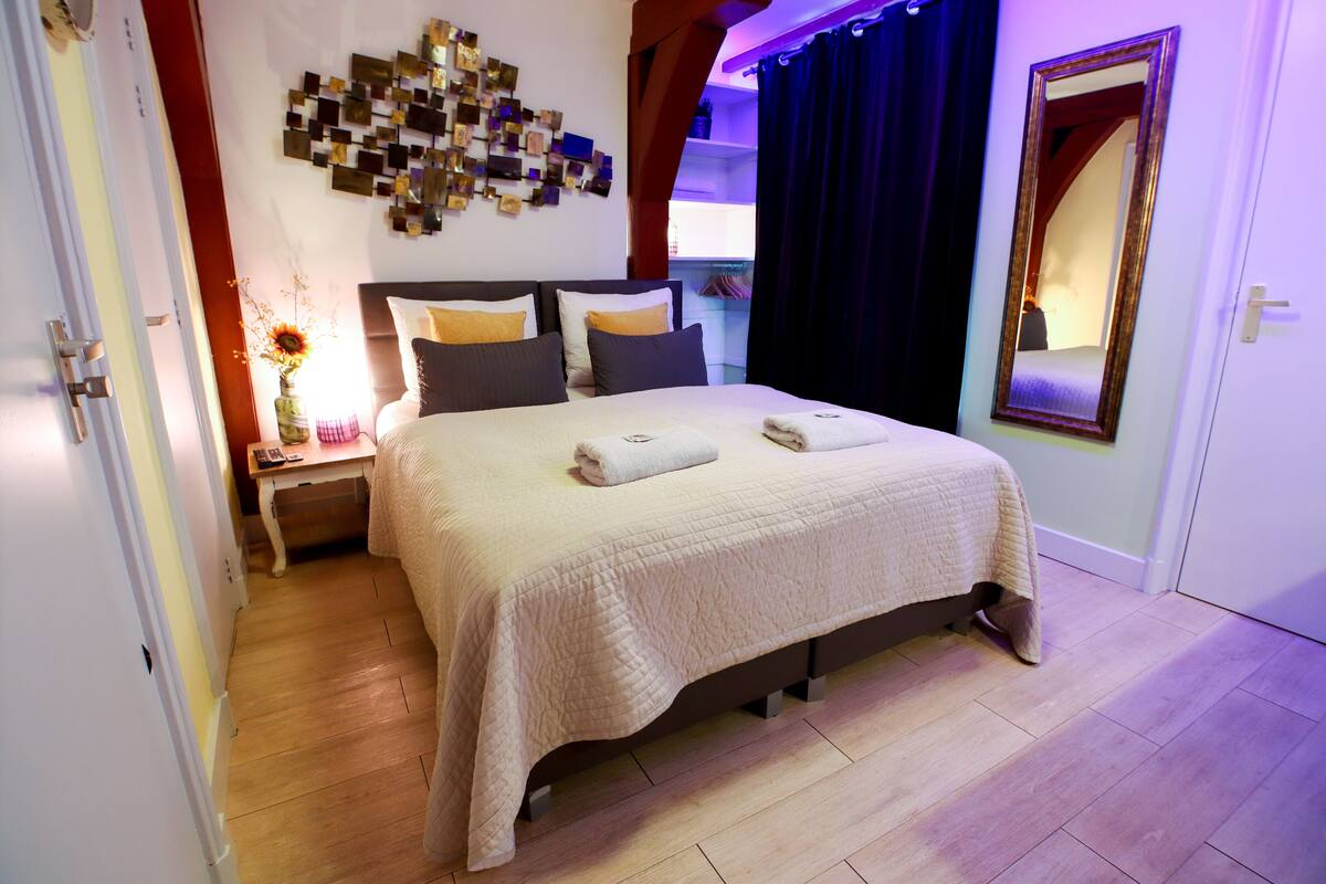 Cama bem arrumada com edredom branco e almofadas, mesa de cabeceira com um vaso de flores e uma obra de arte na parede acima da cama.