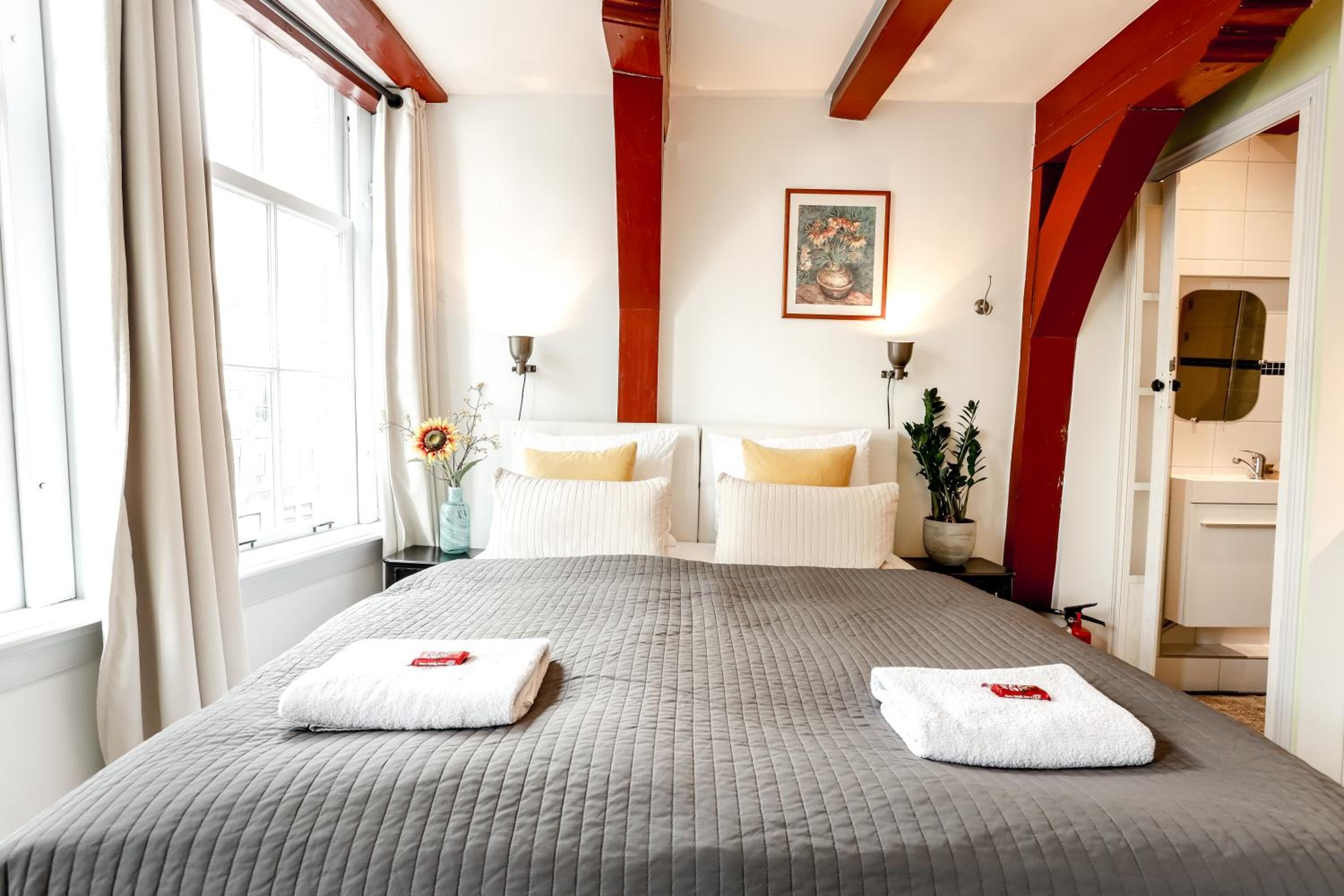 A hotel room with a large bed, two bedside tables, a painting on the wall, and a bathroom visible through an open door.