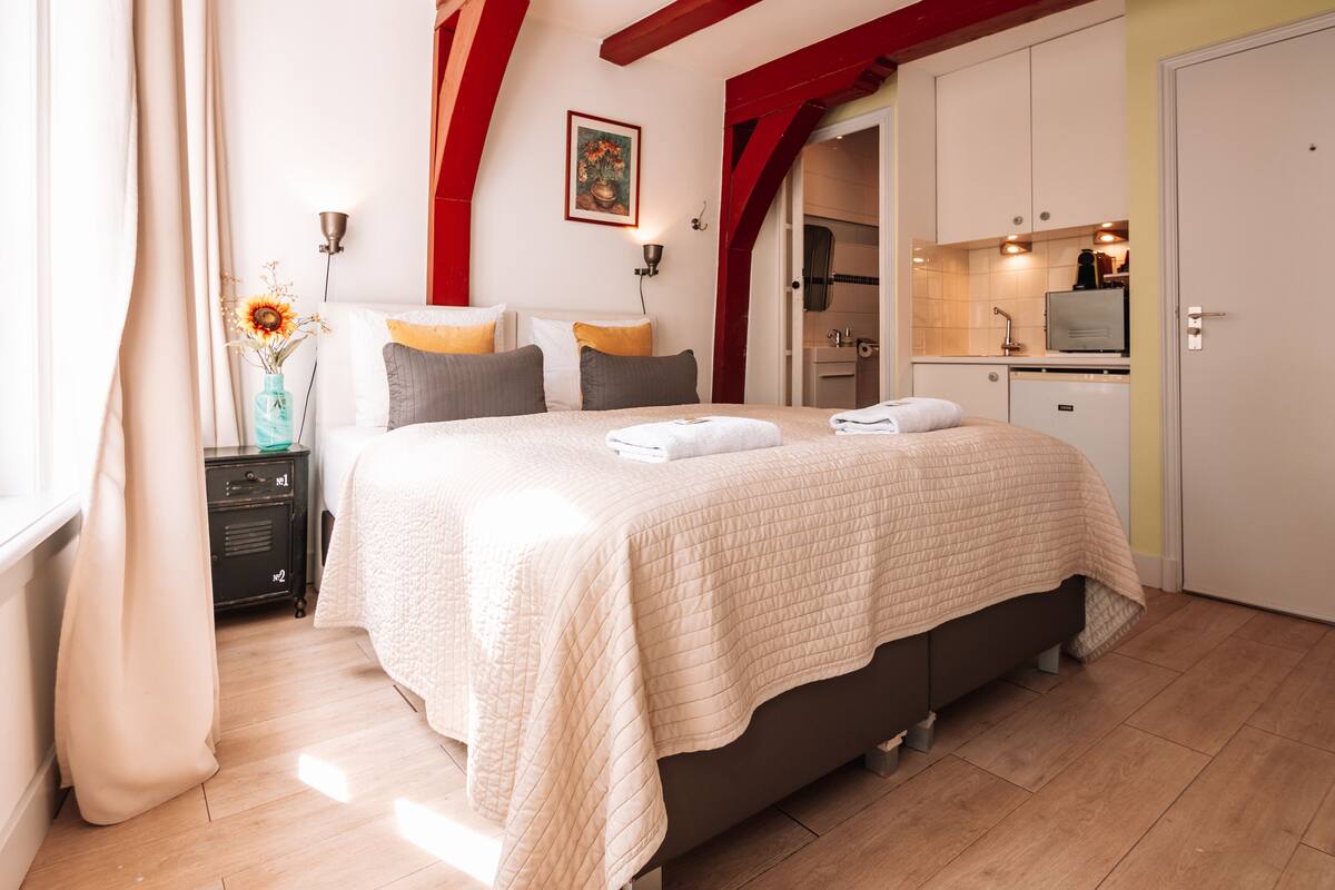 Quarto com cama grande, mesa de cabeceira, cortinas na janela e vista para a cozinha.