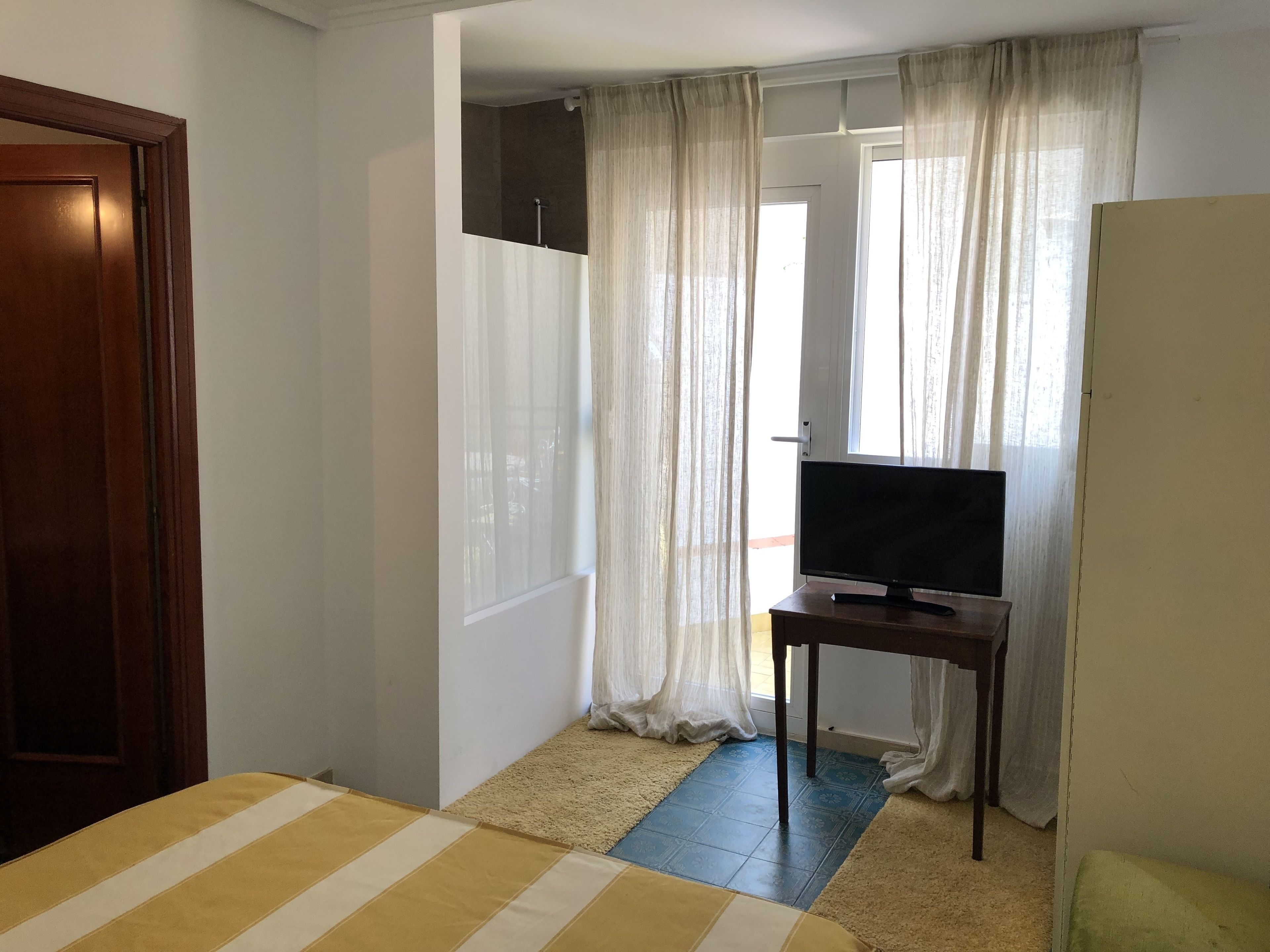 A room with a bed, a television on a small table, and a window with sheer curtains.
