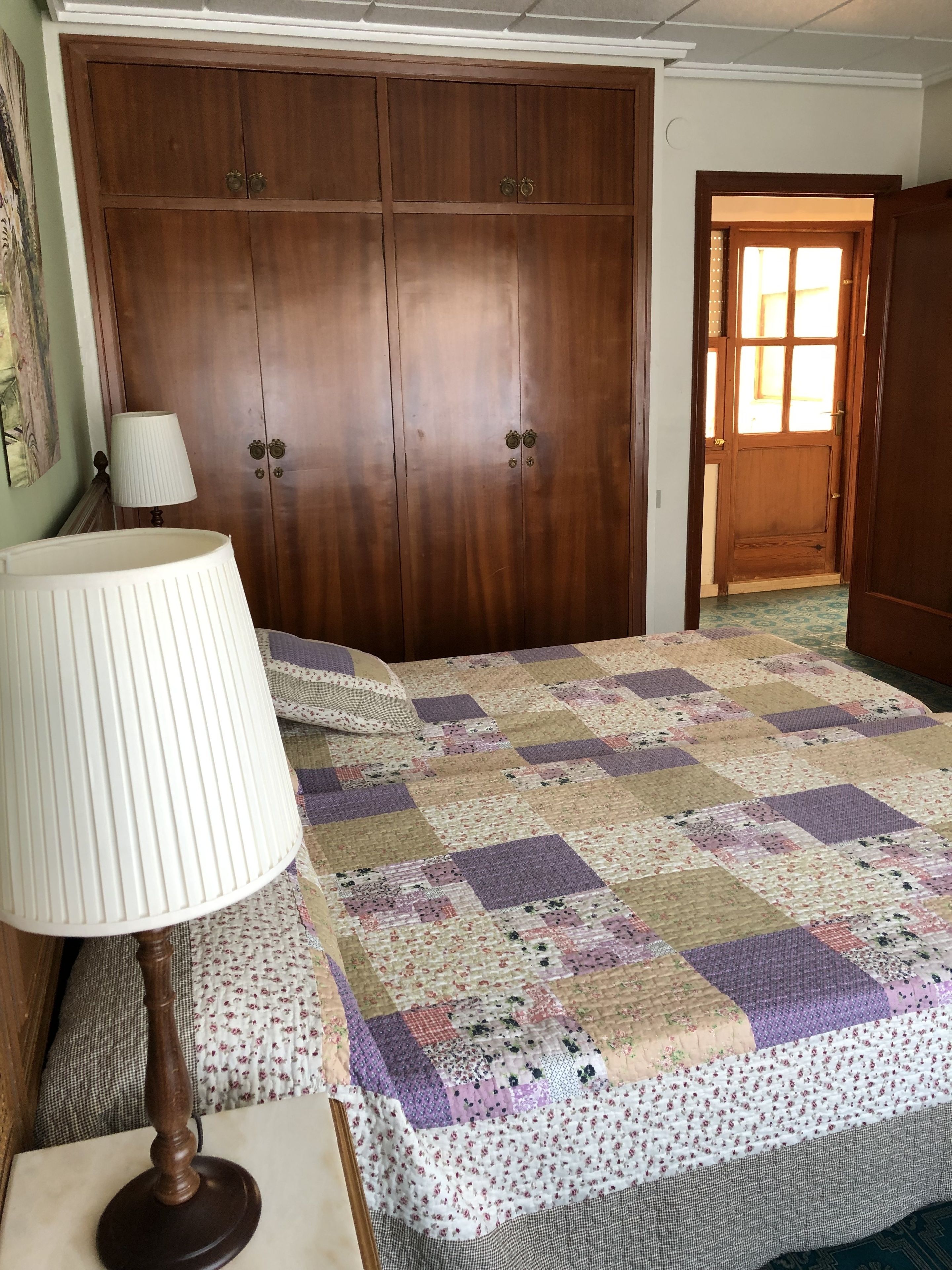 Un dormitorio con un armario de madera, un edredón acolchado, una lámpara de noche y una puerta que da a otra habitación.