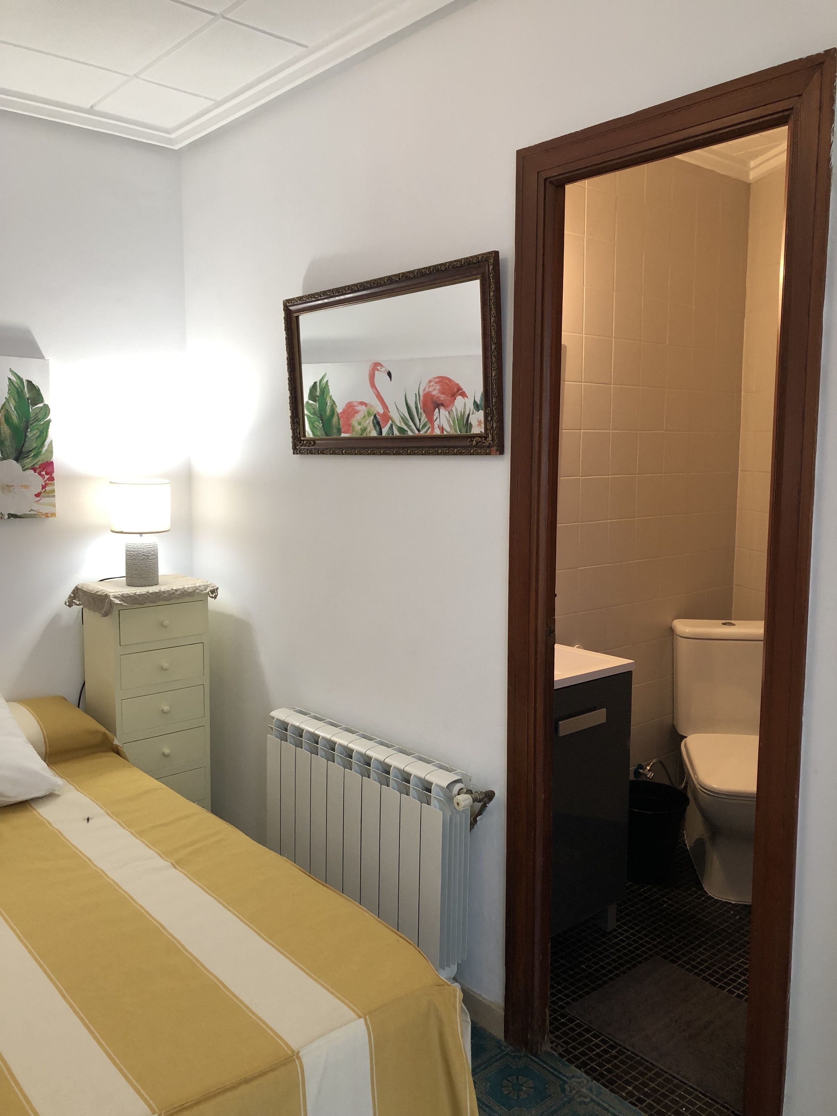 A hotel room with a bed, a radiator, a small cabinet, a mirror with a floral painting, and a view into the bathroom.