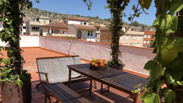 Terraza en la azotea con mesa y banco de madera, rodeada de plantas en macetas y una cesta de fruta.