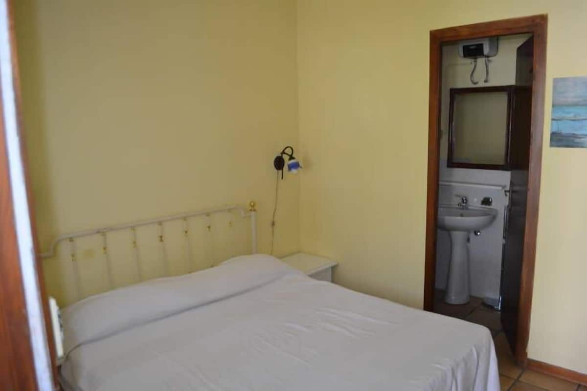Ein kleines Zimmer mit einem Einzelbett, einem Nachttisch, einem an der Wand montierten Fernseher und einem Badezimmer mit Waschbecken und Spiegel.