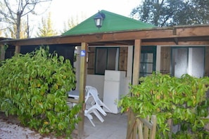 Un edificio pequeño de una sola planta con techo verde y un patio cubierto. Se ve un refrigerador blanco y una silla de plástico.