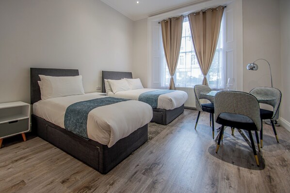 A hotel room with two beds, a small table, and a chair. - New Kent Apartments (London)