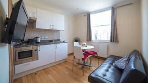 A compact kitchen with modern appliances, a dining area with a table and chairs, and a leather sofa in a bright room with a large window. - New Kent Apartments (London)