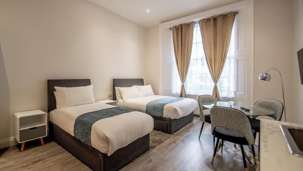 A hotel room with two beds, a small table, a chair, and a desk with a lamp. - New Kent Apartments (London)