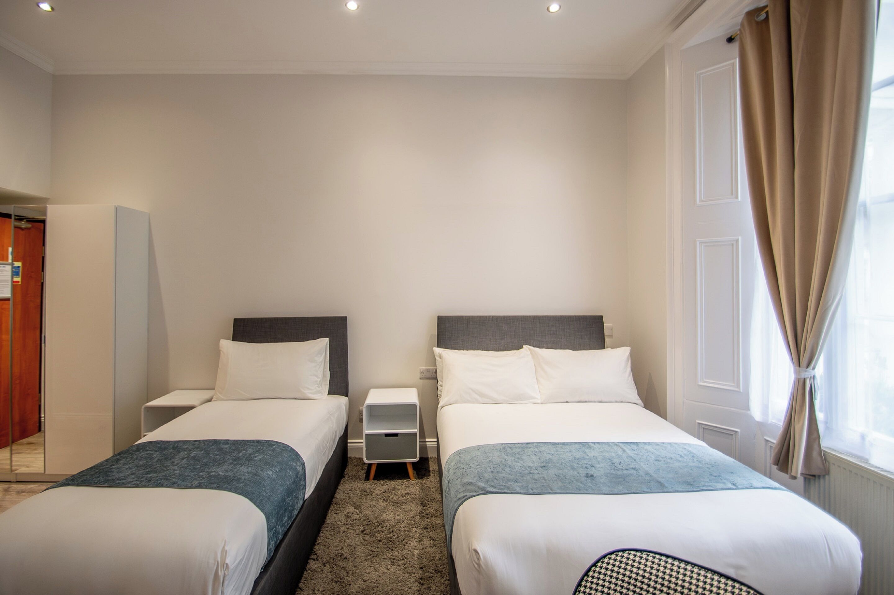 Ein Hotelzimmer mit zwei Betten, einem kleinen Nachttisch, einem Beistelltisch und einem Fenster mit Vorhängen.