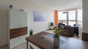 A modern room with a wooden desk, a white wardrobe, a painting of a coastal scene, and a balcony with orange curtains. - Strandhaus Christiansen (Cuxhaven)