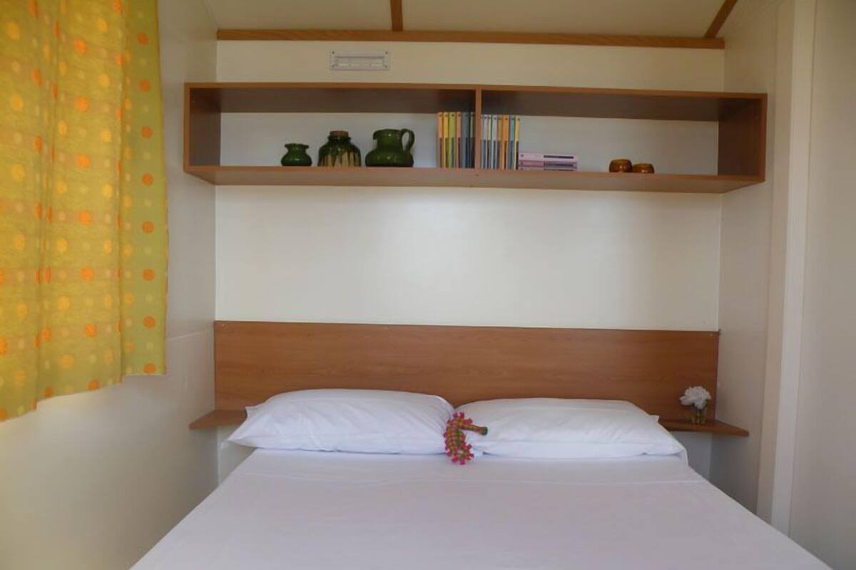 Un letto singolo con lenzuola bianche, una testiera in legno e uno scaffale sopra con libri e oggetti decorativi.