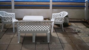 Un patio con dos sillas blancas y una mesa, con vista a una playa, el océano y un cielo despejado.
