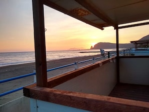 Vista a la playa desde un área techada con barandilla, una terraza de madera y el ocaso sobre el océano.