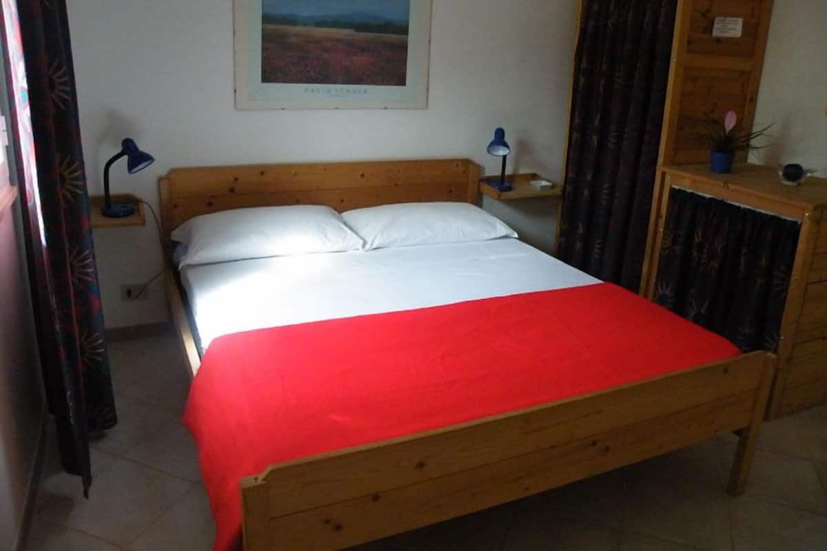 Una camera da letto con un letto in legno, biancheria bianca, una coperta rossa, un comodino con una lampada, un'armadio in legno e un quadro incorniciato appeso al muro.