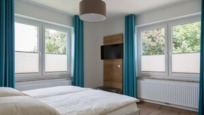 Une chambre à coucher avec un lit, un téléviseur fixé sur un panneau en bois et de grandes fenêtres équipées de stores.