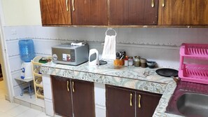 A kitchen with wooden cabinets, a microwave, a kettle, a water dispenser, and a pink dish rack.