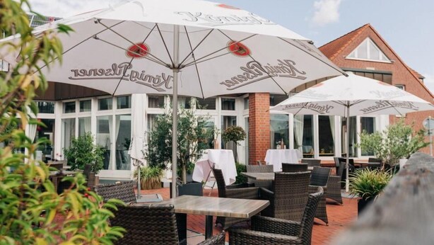 An outdoor seating area with wicker furniture and white umbrellas with a logo. - Merfelder Hof (Dülmen)