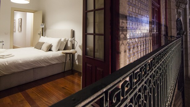 Quarto com cama, mesa de cabeceira e vista para um corredor com paredes estampadas e corrimão.