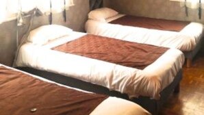 A room with three single beds arranged in a row, each with a white mattress and a brown blanket. There is a window on the left side of the room, and a door is visible on the right. - Le Petit Blanzou (Pierre-Buffiere)