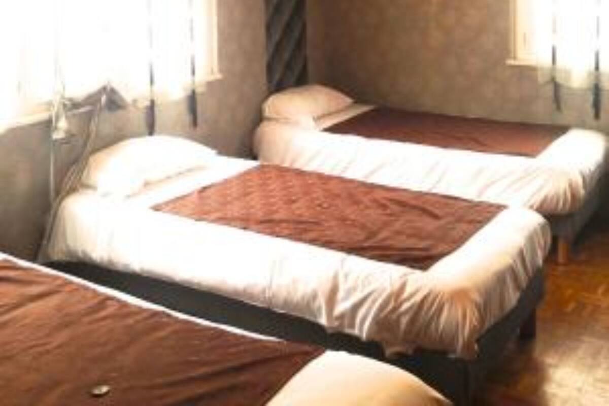 A room with three single beds arranged in a row, each with a white mattress and a brown blanket. There is a window on the left side of the room, and a door is visible on the right.