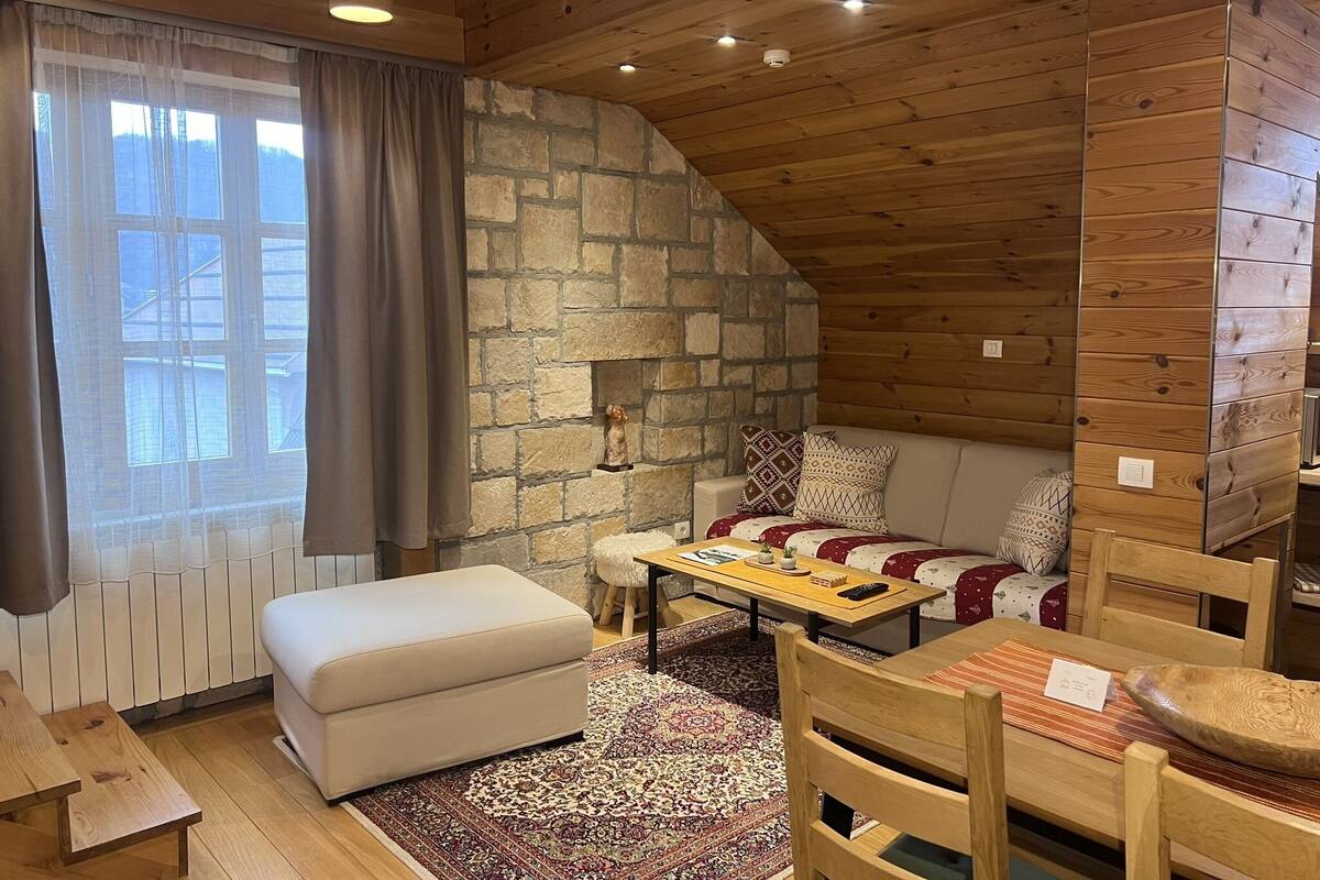 Une pièce chaleureuse avec un mur en pierre, un plafond en bois, un canapé, une table et une fenêtre avec des rideaux.
