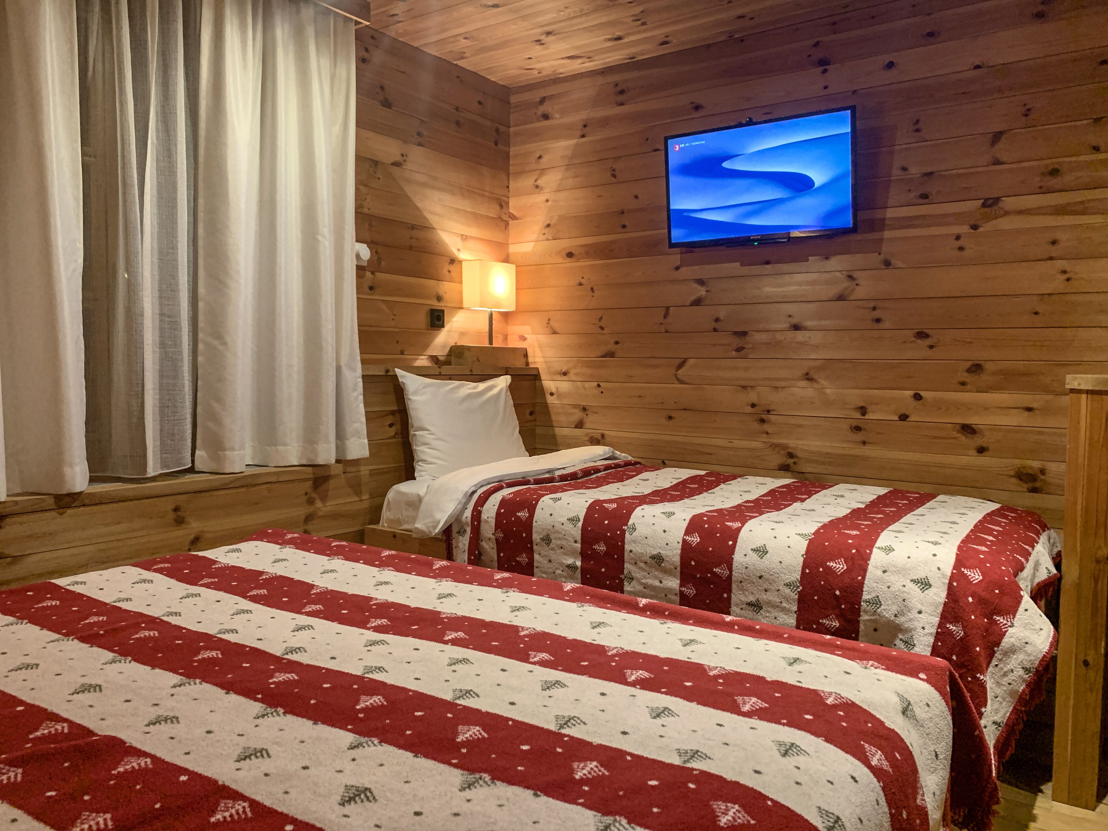 A cozy cabin room with wooden walls, a bed with a red and white striped blanket, a television on the wall, and a window with white curtains.