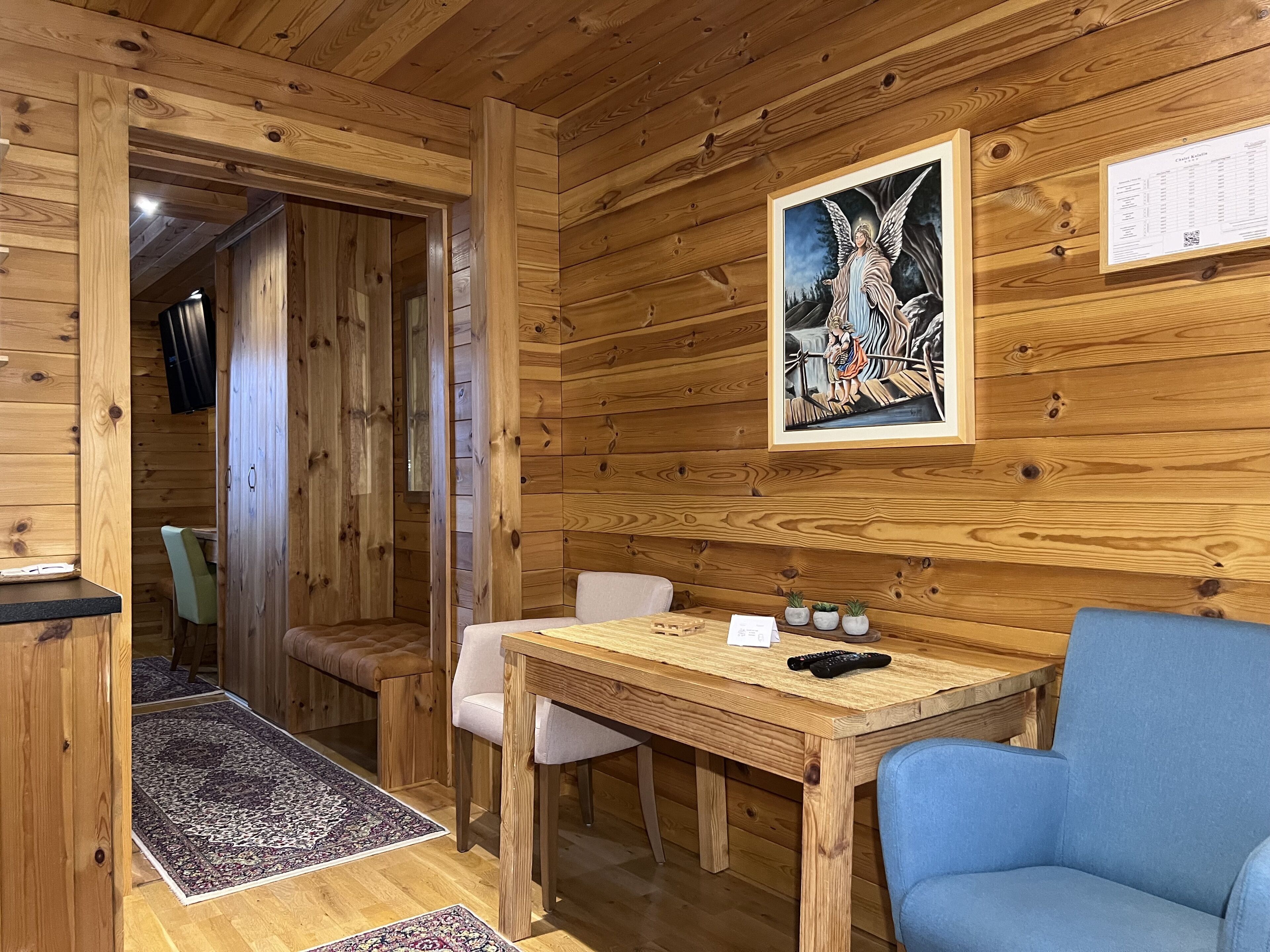 Innenansicht einer Holzhütte mit Essbereich, einem blauen Stuhl, einem Tisch mit Fernbedienung und einem gerahmten Bild an der Wand.