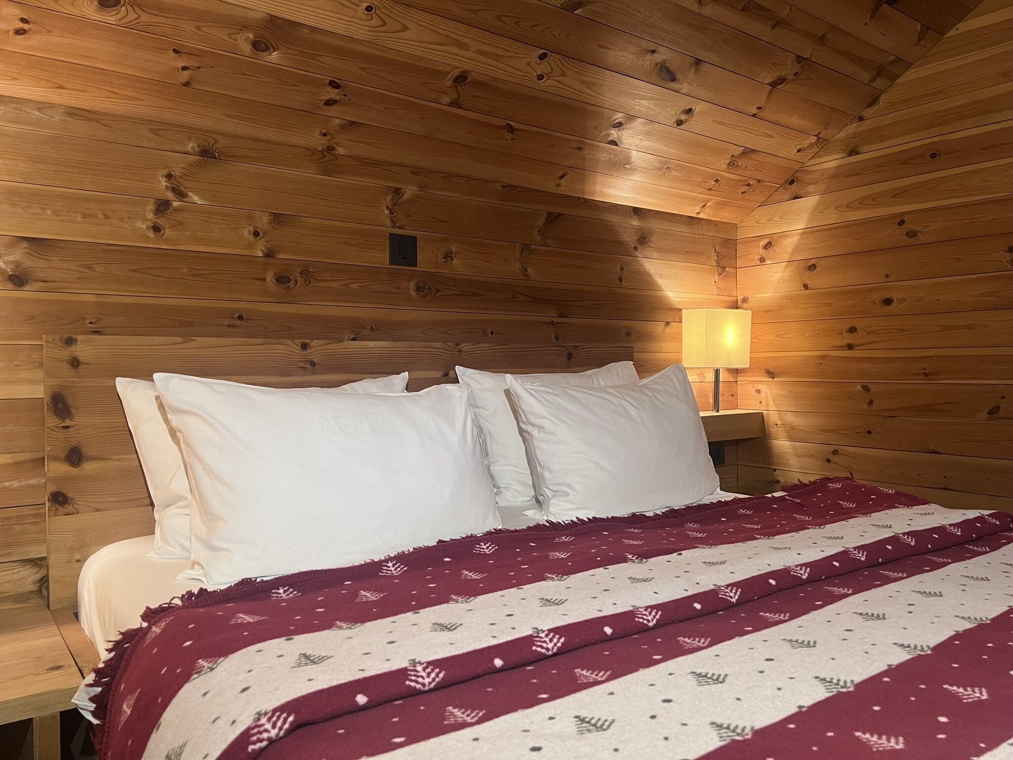 A wooden cabin-style room with a bed, white pillows, a red and white patterned blanket, and a nightstand with a lamp.