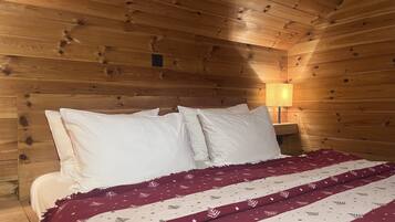 A wooden cabin-style room with a bed, white pillows, a red and white patterned blanket, and a nightstand with a lamp.