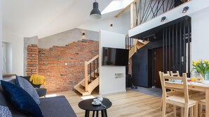 Ein moderner Wohnraum mit Backsteinwand, einer Treppe, einem Flachbildfernseher, einem Essbereich mit Holzmöbeln und einer Couch mit Kissen.