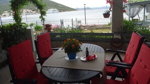 Una terraza con una mesa para dos, sillas rojas, una planta en maceta con flores amarillas y una vista al agua y colinas en el fondo.