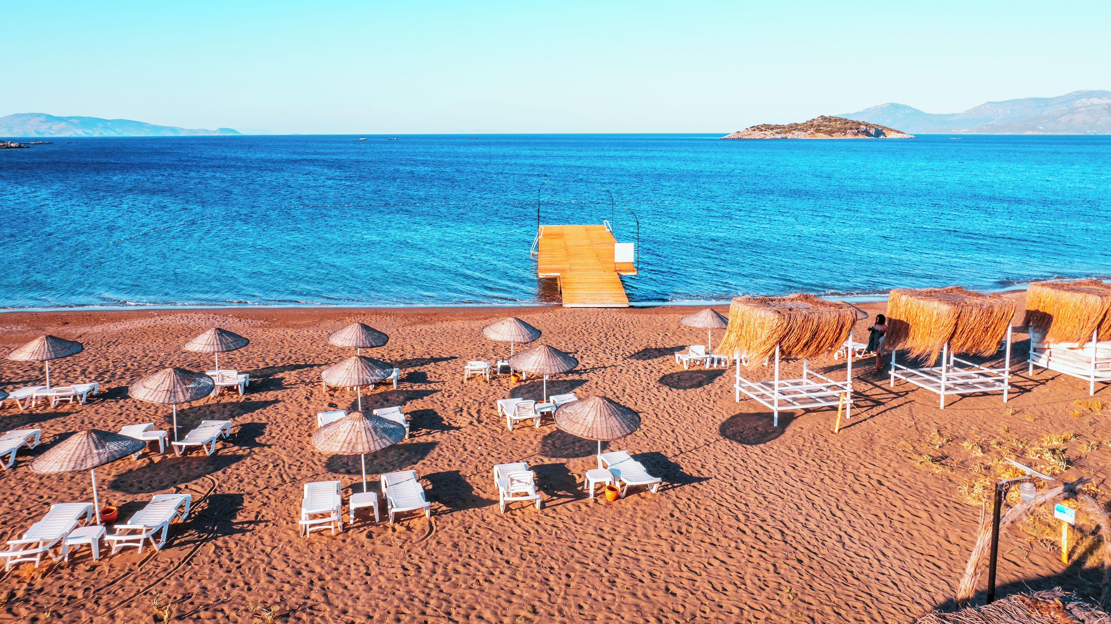 Une plage avec des chaises longues blanches et des parasols en paille, une terrasse en bois et une vue dégagée sur la mer et les îles lointaines.