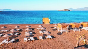 Une plage avec des chaises longues blanches et des parasols en paille, une terrasse en bois et une vue dégagée sur la mer et les îles lointaines.