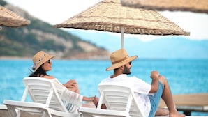 Two people relaxing on white lounge chairs by a pool with a view of the sea and hills in the background. -  Acroter Hotel & Spa Datca (Datça)