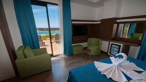 A hotel room with a bed, a TV, a green sofa, a small table, and a view of a pool and mountains. -  Acroter Hotel & Spa Datca (Datça)