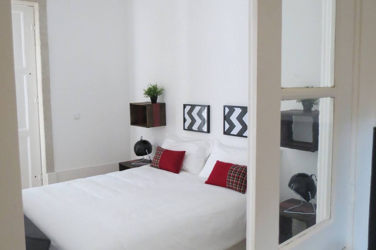 A bedroom with a bed, pillows, a nightstand, a plant, and wall decorations.
